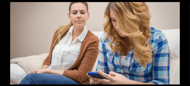 Parent looks suspiciously over daughter's shoulder as daughter looks at her phone.